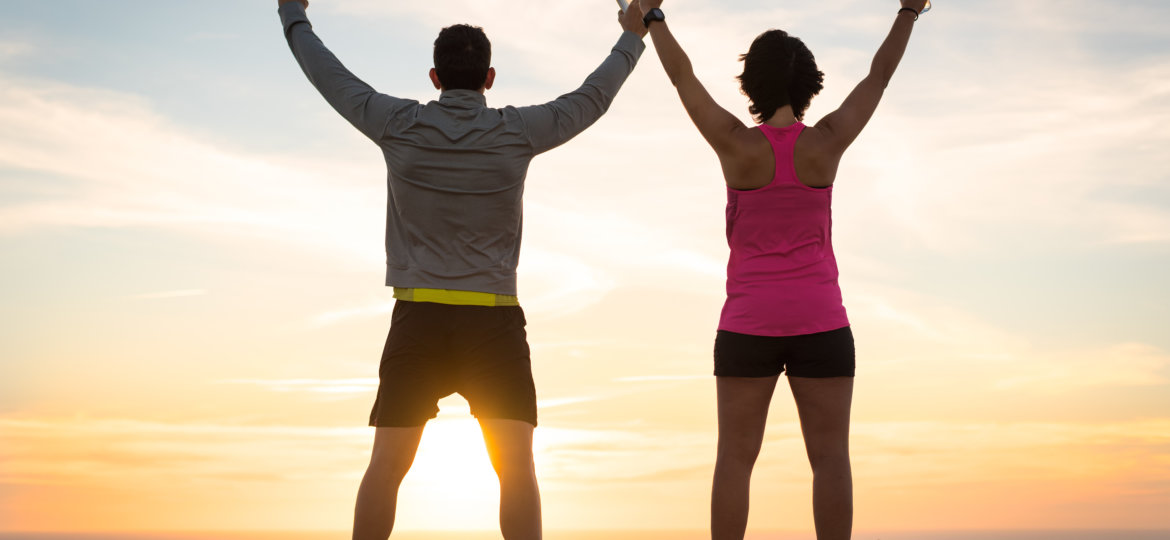 Sporty runner couple rising arms in victory sign after successful training outdoor on the coast facing the sun showing back. Running team success concept.
